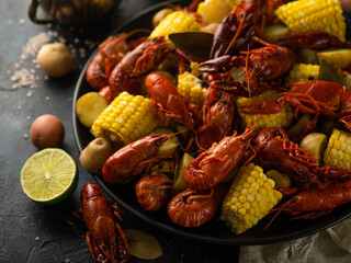 Boiled corn, crayfish on a large black plate. Also on the table we see lemon, cancer, potatoes. Close-up. Careful viewing. Bright colors. No people. Dark background.