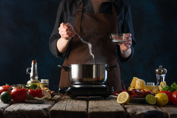 The photo shows the process of making vegetable soup. There is a saucepan on a wooden table, and many ingredients are laid out on the table. The cook adds spices to the soup. Blue background.