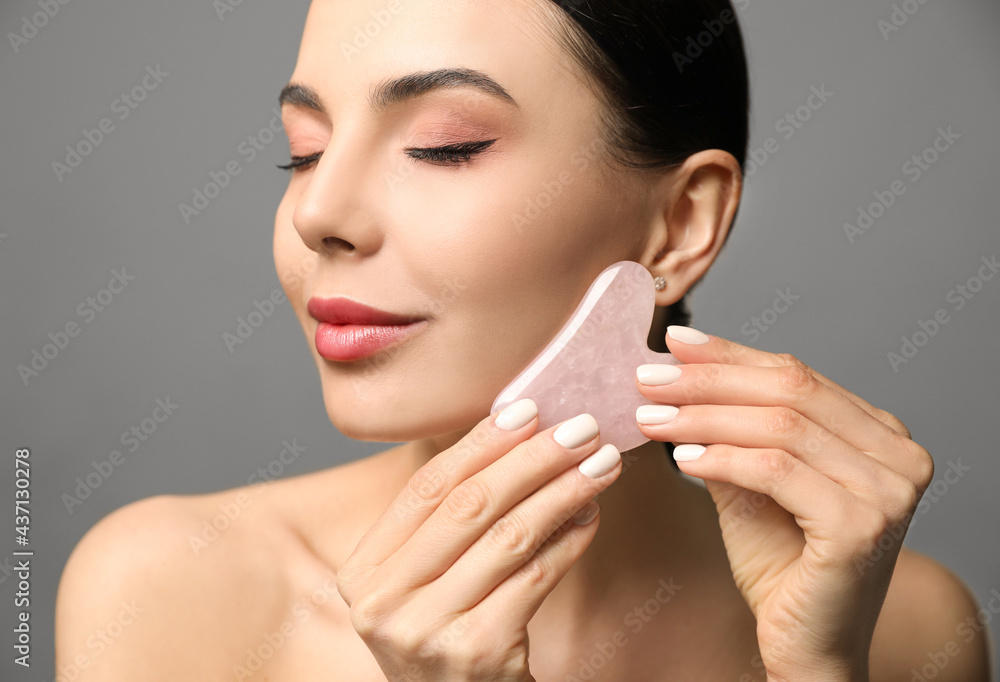 Poster Beautiful young woman doing facial massage with gua sha tool on grey background, closeup