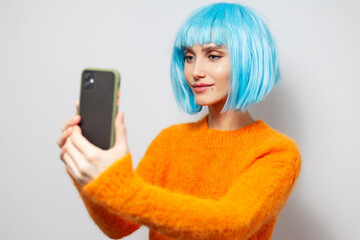 Studio portrait of young pretty girl, smiling and taking selfie photo by smartphone on background of white; wearing orange sweater and blue hair bob.