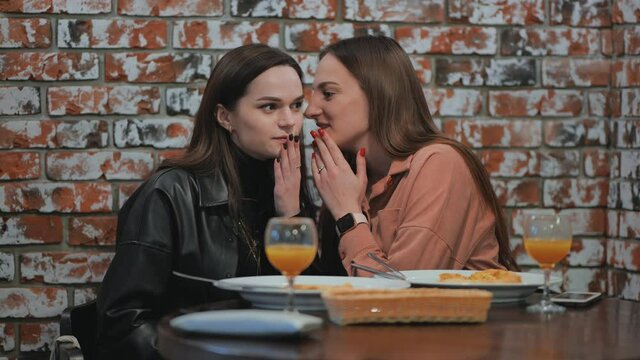 Young and cheerful girls gossip and whisper to each other while sitting in a cafe.