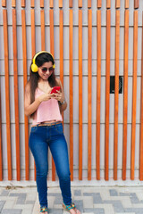Cheerful young woman with cool glasses and fashionable clothes walking down the street enjoying playlist songs on headphones and reading good news sms on smartphone