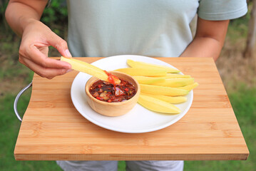 Hand dipping mango slice in sweet fish sauce on white plate over bamboo wooden board background