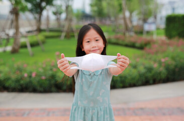 Little Asian girl child showing face mask in the public garden during coronavirus outbreak. Focus at mask in his hands