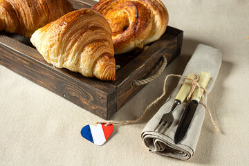 Fresh french croissants in a wooden tray, a heart in the colors of the French flag and cutlery on a linen tablecloth