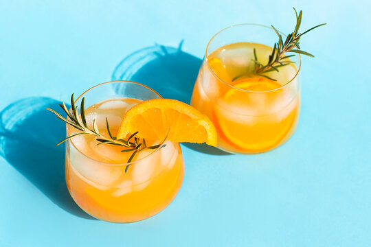 Lemonade, orange cocktail, refreshing summer drink with ice and orange slices in a glass with a sprig of rosemary on a blue background on a bright sunny day
