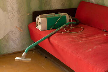Old red couch and a vintage vacuum cleaner in a unclean room.