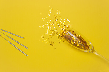 Confetti stars spill out of a champagne glass and a sparkler stick on a yellow background. The view from the top. The concept of the celebration.