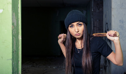 Fototapeta premium A fighter girl, a brunette in a black hat, stands in an old building. With two hands, he holds an ancient Chinese weapon - nunchucks - on his shoulders. Looks into the camera. Complete composure 