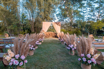 A rustic garden wedding setup, the main aisle lined with baskets with pampas leaves and pastel pink...