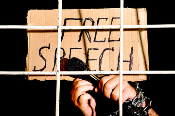 the concept of human rights freedom of speech hands shackled with an iron chain hold a microphone and a cardboard sign with the inscription free speech behind an iron prison bars