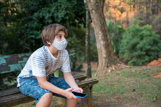 garoto usando m&aacute;scara de prote&ccedil;&atilde;o contra covid19, sentado no bando do parque, pensando e refletindo. 