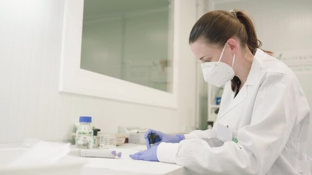Woman Wearing Mask And Laboratory GownSitting On Table And Extracting Plant Sap To Analyze Viruses In Biotechnology Laboratory. - medium shot