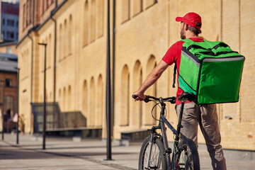 Rear view of delivery man standing with bicycle and thermo bag on the sunny street