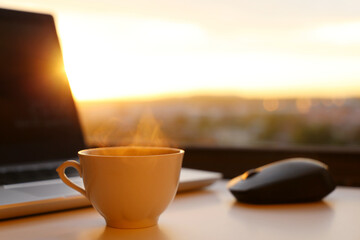 Hot coffee cup and laptop on table against the window at sunset. Cozy workplace in home office, concept of remote work and earning online