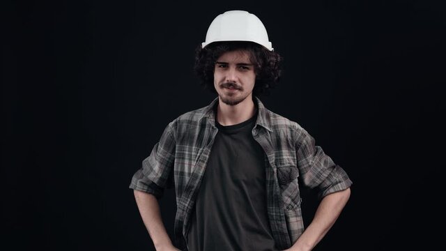 The charismatic hipster boy puts on his engineer's helmet and looks proudly at the camera. Isolated on black background. The concept of life. People's emotions. 4k portrait