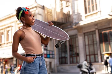 Rolgordijnen Portrait of happy african-american woman with skateboard. Young stylish woman with skateboard outdoors.. © JustLife