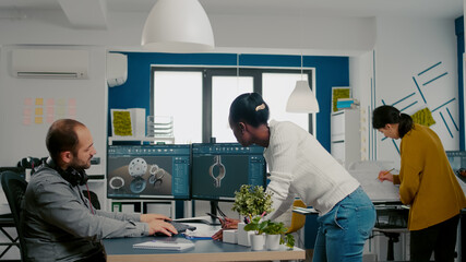 Multiethnic team sharing ideas about industrial project using two monitors with 3D gears and metalic clamp using CAD software. Industrial engineer and black manager innovating prototype of motor