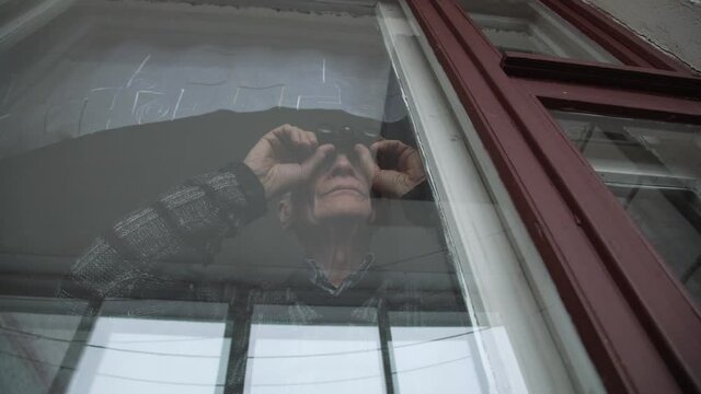 Crazy aged male person holds binoculars in hands and watching through window indoor of nursing house