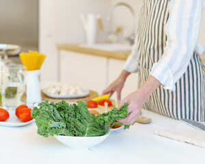 Cropped shot of retired woman in apron preparing fresh healthy vegetable salad while cooking food for whole family during retirement in modern light kitchen at home, fresh organic vegetables on table