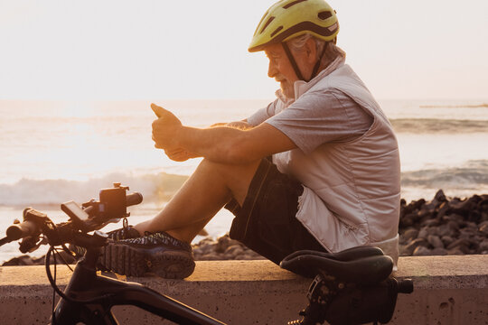 Cyclist man using mobile phone sitting along the sea at sunset, close his bike. Retired people enjoying healthy lifestyle