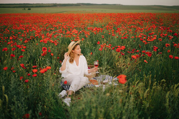 Naklejka na ściany i meble Young beautiful woman wearing white linen suit and straw hat having picnic with pizza, strawberries and wine on a poppy field.