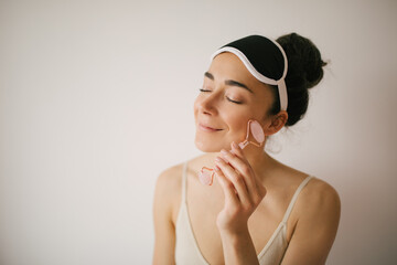 Beautiful young woman with perfect skin wearing sleep mask using a jade face roller with natural...