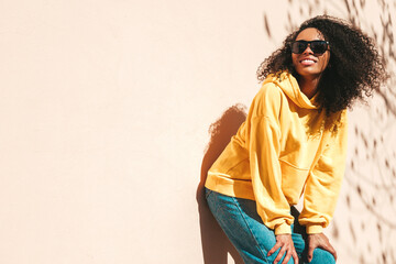 Fototapeta premium Beautiful black woman with afro curls hairstyle.Smiling hipster model in yellow hoodie. Sexy carefree female posing on the street background in sunglasses. Cheerful and happy