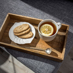 A white cup of tea with lemon, a long cupronickel spoon with a twisted handle and a saucer with three oatmeal cookies for breakfast on a wooden tray, a rag napkin on a gray sofa.