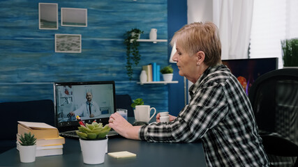 Remote young physician using webcam prescribing medicine to sick aged woman sitting in living room via internet. Patient talks with therapist by videoconference call application, holding pills flask