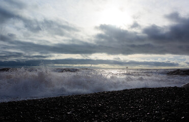 sun shining through clouds with a pebble beach in the foreground and waves breaking on the shore