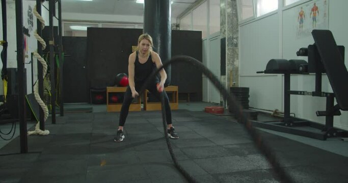 Sport woman doing battle ropes exercise at gym. Fitness girl athlete with fit body exercising, doing functional training with heavy ropes indoors. Rope workout.