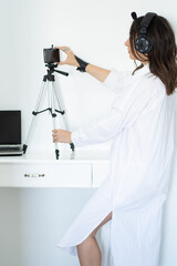 Women in a white long dress is preparing for an online meeting, a conference, a business interview, standing in front of the phone on a tripod. Women with headphones. White fashion concept 