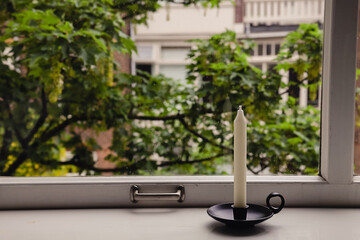 Long white candle in an old style vintage candle holder standing  on the wooden big window sill on a blurred green trees backgroung. Slow life and cottage core concept.