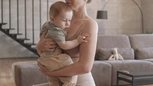 Tilt up of handicapped good-looking sporty woman with leg prosthesis sitting on fitball at home, holding little son, hugging and kissing him
