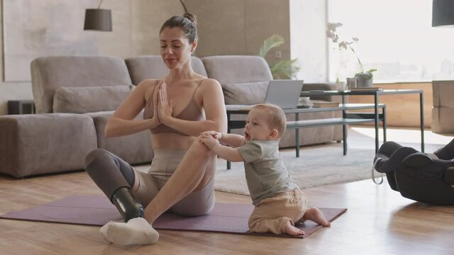 Lockdown of disabled female yogi wearing stylish sportswear with leg prosthesis meditating on rubber mat in apartment while cute toddler sitting nearby