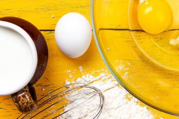 Pancake ingredients. Flour, chicken eggs, milk and handmade whisk on a yellow background. Preparing...