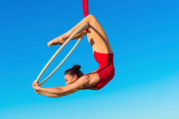 acrobat athletic, young graceful gymnast performing aerial exercise in the air ring outdoors on sky background. flexible woman in red suit performs circus artist dancing on hoop demonstrates poses. - obrazy, fototapety, plakaty