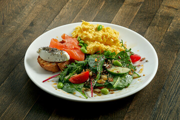 Delicious and hearty breakfast - scrambled eggs and toast with cream cheese and salmon, mix salad in a white plate on a wooden background