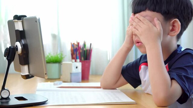 Cute tired Asian elementary student kid yawning and rubbing eyes strain while studying online lessons via computer tablet on his desk at home. Online learning technology in education and mental health
