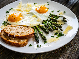 Tasty breakfast. Sunny side up eggs with green asparagus and parmesan served on black plate on wooden table
