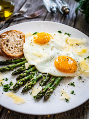 Tasty breakfast. Sunny side up eggs with green asparagus and parmesan served on black plate on wooden table
