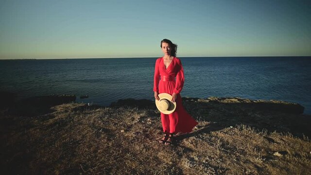 An attractive young Caucasian woman in a red dress with long hair stands with a straw hat in her hands on a rocky seashore with closed eyes in windy weather. Shallow depth of field