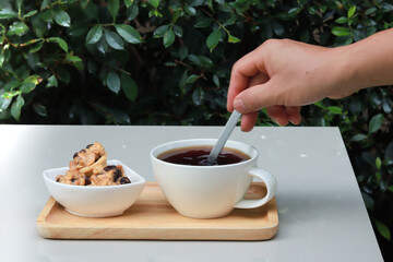 Coffee and cookies served on a wooden tray, on a white table, and a hand holding a spoon to stir coffee.  Garden atmosphere. The background is blurred green leaves.