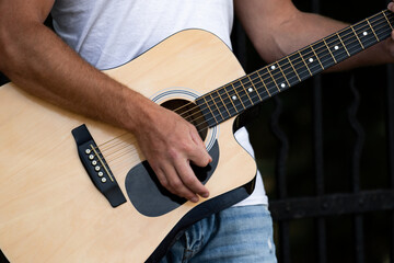A street musician guitarist stands and grazes in a park on alley avenue in the city on the street near the fence shot close-up and focus on the guitar and hands in blue jeans and a white T-shirt.