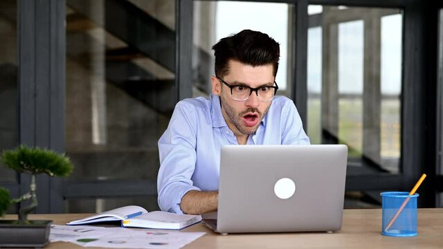 Annoyed caucasian office employee, freelancer or broker sitting at a table in a modern office, indignantly looking at a laptop, overworked, stressed, gesturing with his hands, shouting