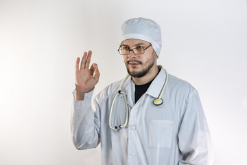 A young, dark-haired man with a beard, wearing a white medical coat and a beanie, folded his fingers in an approving gesture against the light background.