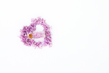 Glass bottle standing on a white background surrounded by lilac flower petals in the shape of a heart. Idea for floral fragrances.