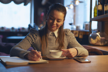 Beautiful woman sitting in a cafe at a table drinking tea and making notes in a notebook