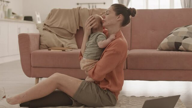 Wide shot of mid adult disabled Caucasian woman with artificial leg sitting on floor on carpet at home, holding toddler in hands while trying to work on laptop